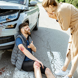 An injured man sits next to a crashed car while a woman checks on him after an accident