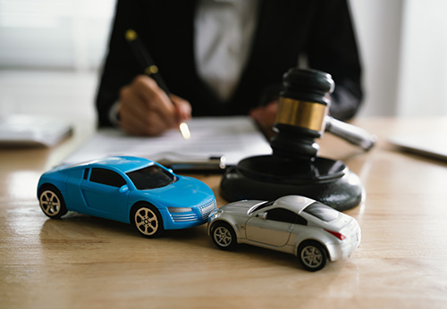 Lawyer signing documents with two toy cars and a gavel on the desk.